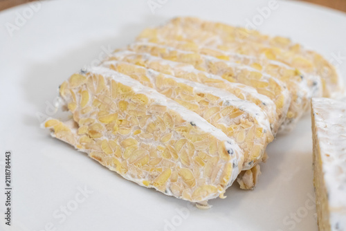 Sliced raw organic tempeh block on a clean white plate. Close up view showing the rich texture of fermented soybeans bound together by white mycelium. A healthy, plant-based vegan protein