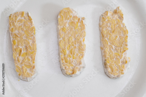 Sliced raw organic tempeh block on a clean white plate. Close up view showing the rich texture of fermented soybeans bound together by white mycelium. A healthy, plant-based vegan protein