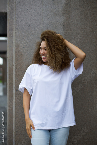 Smiling young woman with curly hair posing casually in white t shirt and light jeans beside textured stone wall outdoors with relaxed urban style and natural confidence