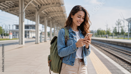 A young woman with a backpack smiling while using her smartphone at an empty train station platform on a sunny day.