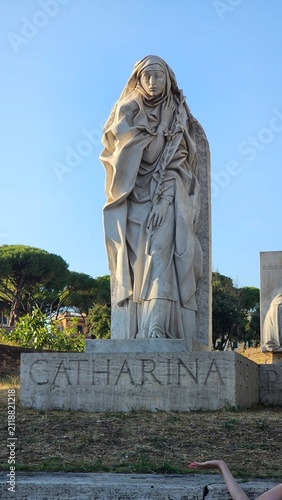 Принти на полотні A marble statue of Saint Catharina stands on a stone pedestal with her name inscribed, surrounded by trees under a clear blue sky
