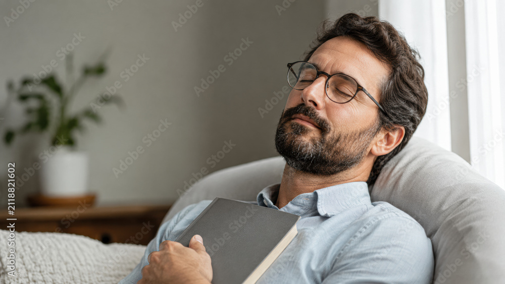 Fototapeta premium Man resting on a couch with closed eyes, holding a book, appearing relaxed and peaceful in a cozy indoor setting.