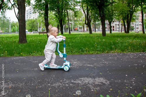 A young child moves quickly on a scooter while enjoying a day in the park surrounded by green grass and trees. The area is lively with other buildings nearby.