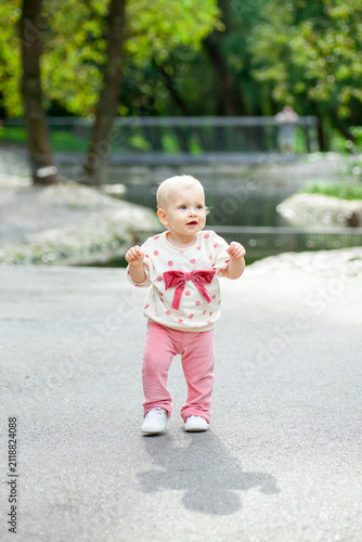 A young child walks carefully on a path in a park. Trees and a small body of water are nearby. The child looks happy while trying to balance on their feet.