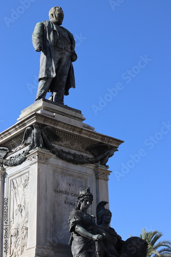 Bild auf Leinwand Bronze statue of Camillo Cavour stands atop an ornate stone pedestal with allegorical figures beneath a clear blue sky