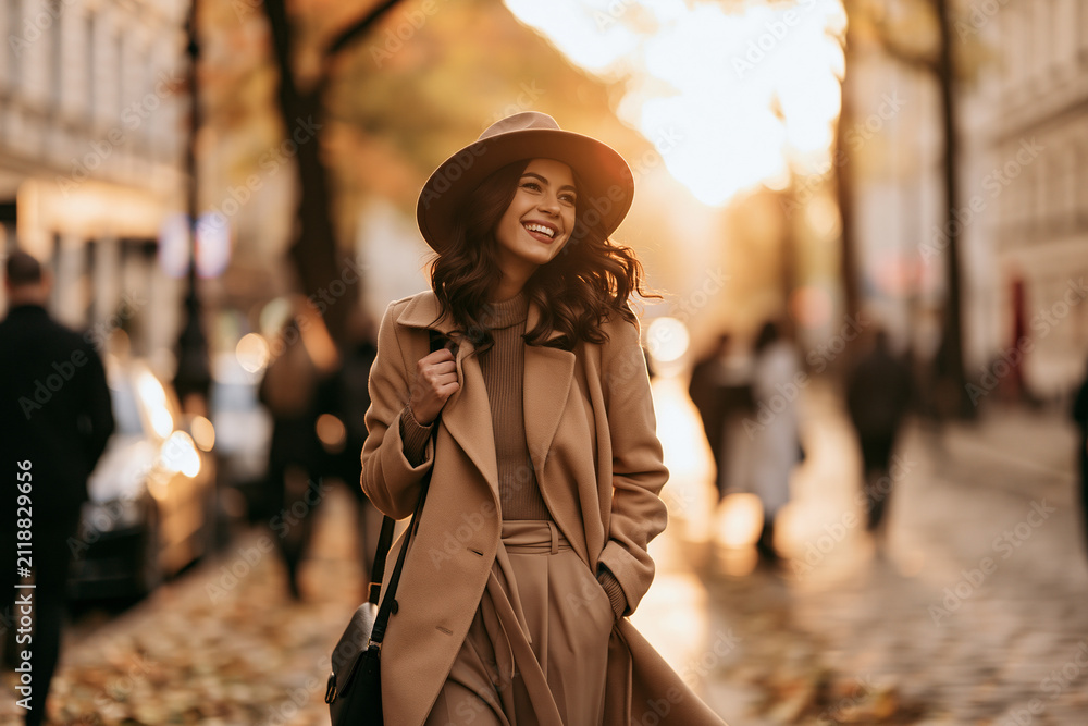 Fototapeta premium A stylish woman in a tan coat and wide-brimmed hat walks happily down a sun-drenched city street during a beautiful autumn afternoon
