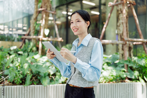 Smiling businesswoman holding a tablet and stylus in a green modern office garden, representing digital work, sustainability, productivity, leadership, career growth, and professional success.