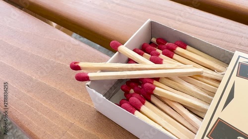 Close-up of open cardboard matchbox filled with wooden matches with red tips, resting on a wooden surface. A few matches are scattered outside the box. Soft natural light and subtle camera movement