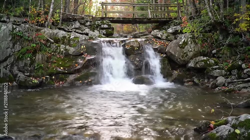 A serene forest waterfall flows over mossy rocks beneath a rustic wooden bridge in warm sunlight