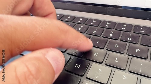 Detailed close-up of a hand typing on a black laptop keyboard covered with dust and small particles. Fingers press the keys in a natural typing motion. Shallow depth of field focuses on the dusty keys