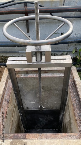 Close-up of a stainless steel manual sluice gate valve installed in a wastewater treatment facility. Industrial flow control equipment with handwheel mechanism used for regulating water level, drainag