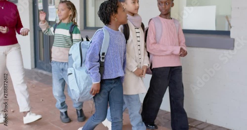 Diverse female pupils walking school path with teacher gesturing, girls stopping to view bulletin
