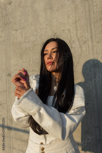 Fotografie Close-up portrait of a thoughtful young Asian woman with long dark hair posing against a textured concrete wall in warm sunlight, wearing a trendy white denim jacket
