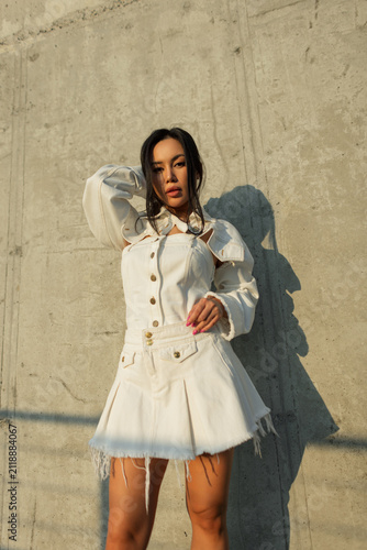 Foto Medium shot of a beautiful young Asian woman in a trendy white denim outfit posing confidently against a textured concrete wall under bright sunlight