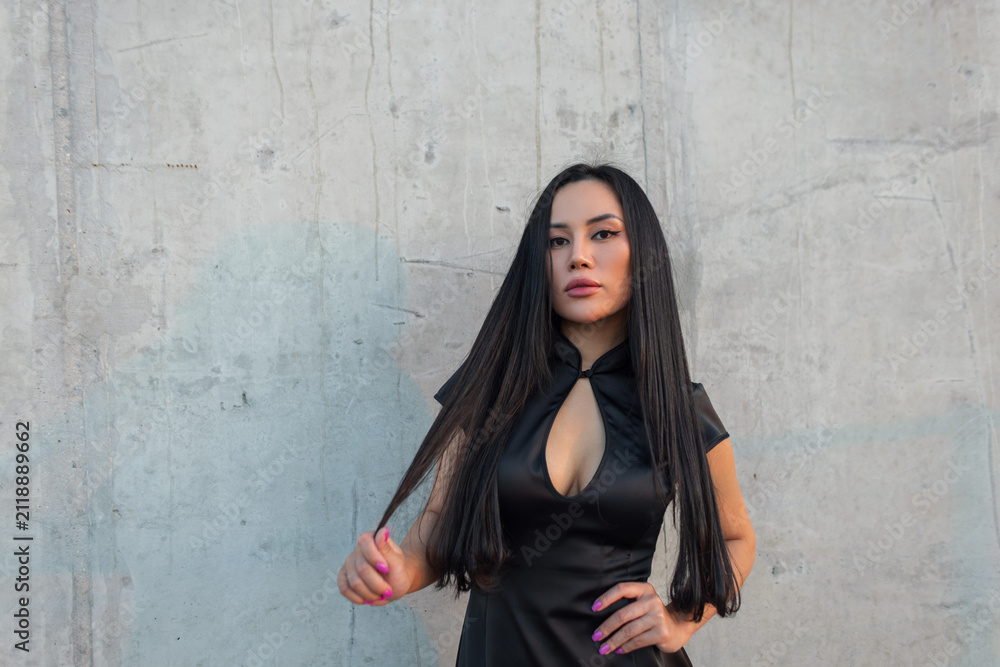 Fototapeta premium Confident Asian woman with long black hair, wearing a modern black cheongsam dress, looking at camera against a textured concrete wall, hand on hip. Pretty glamour urban elegant girl