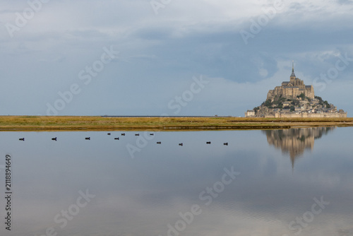Obraz na plátně Scenic wide shot of Mont Saint-Michel on the horizon with swimming ducks creating ripples across mirror-like tidal waters under an overcast sky in France
