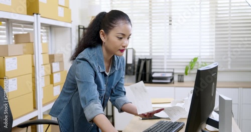 Asian woman small business owner pulls printed shipping label from desk printer, checks online order details on screen, prepares parcel documentation for e-commerce packaging and delivery process