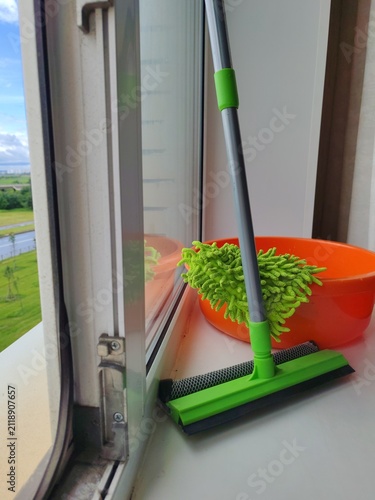 Window cleaning supplies with water bucket,squeegee, green sponge and cloth beside a dirty window in a home interior.