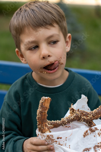 A little boy sticking out his tongue to lick his chocolate-stained lips after eating a sweet pretzel with nuts, sitting outdoors on a bench.