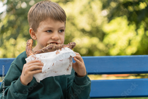 A young boy taking a bite of a chocolate-covered pretzel with nuts while sitting on a blue bench in a park during a sunny day.