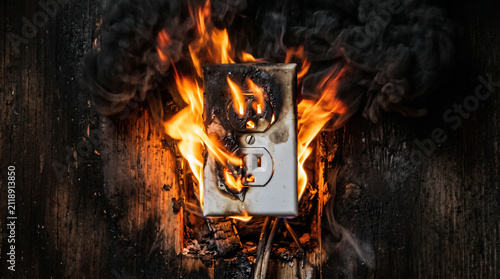 Electrical Outlet Fire Hazard: A severely damaged electrical outlet engulfed in flames, surrounded by thick black smoke and charred debris, highlighting a dangerous fire risk.