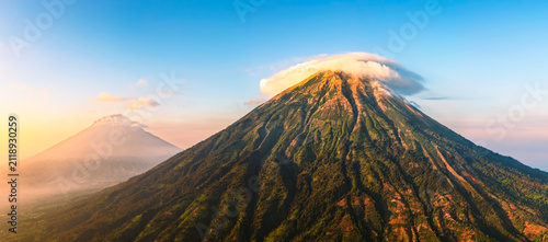 Aerial view of Mount Sindoro with a lenticular cloud cap over its peak, lush green volcanic slopes, and golden hour light in Kejajar, Central Java, Indonesia.