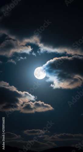 Full moon shining brightly in the night sky surrounded by dramatic, soft clouds and a few visible stars in the darkness