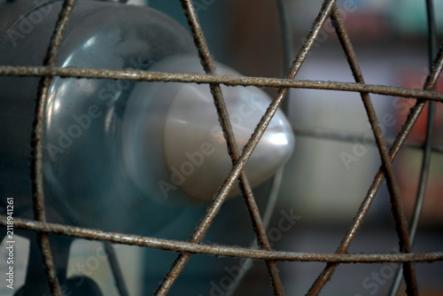 Close-Up Of Vintage Rusty Metal Fan Cage With Blurred Blades And Indoor Background