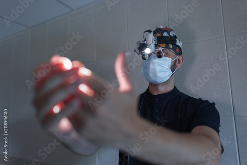 фотографія Focused neurosurgeon wearing a face mask and surgical loupes shines a bright headlamp onto his blurred hands in a clinic room