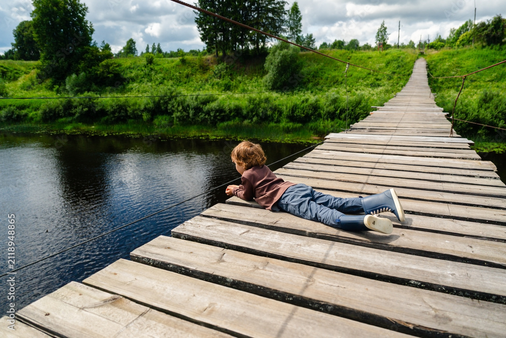 Obraz premium A focused young boy lies on a wooden bridge over a river, patiently fishing while relaxing in nature. This peaceful scene captures a child enjoying a rural retreat and observing the water.