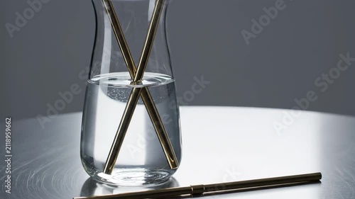 Cool focused light defines a slender glass water carafe with crossed bronze toned reusable straws on a silver table, creating a refined minimalist hydration still life with luminous water reflections