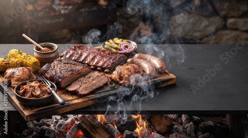 Delicious assortment of smoky grilled barbecue meat featuring ribs, brisket, pulled pork, and sausages on a wooden board over glowing charcoal embers, creating an authentic rustic culinary experience