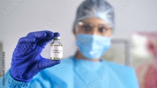 Medical staff wearing personal protective equipment kit showing COVID-19 vaccine. Female doctor in PPE kit holding a new effective vaccine against Coronavirus - flu shot vaccination drive