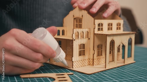 Crafting wooden house model with hands applying glue, installing final wall panel for DIY miniature architecture. New housing concept, creative home construction hobby, handmade real estate prototype