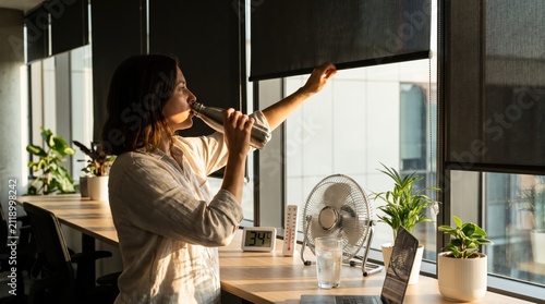 Woman drinks water in hot office during heatwave, practicing safety
