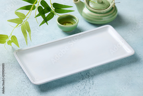 Empty white rectangular plate with green teapot and bamboo leaves. Blue light background. Close up.