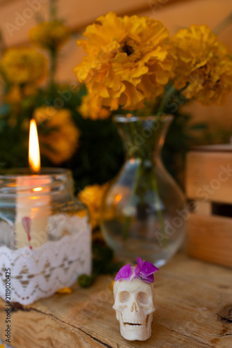 Close-up of decorative skull candle topped with purple flower on rustic wooden table, glowing jar candle with lace wrap and yellow marigolds nearby, soft blurred background and warm festive memorial