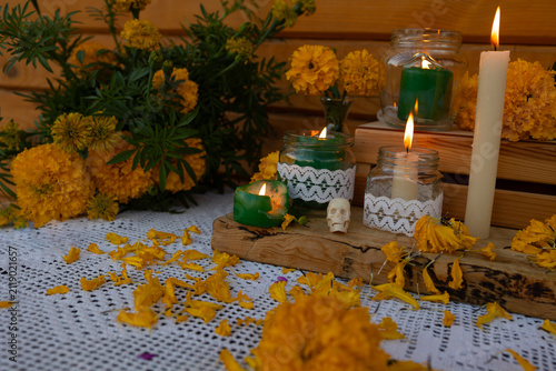 Close view of Day of Dead altar featuring yellow marigold flowers scattered across white lace cloth, lit candles in glass jars, small decorative skull, wooden accents, and warm ritual atmosphere