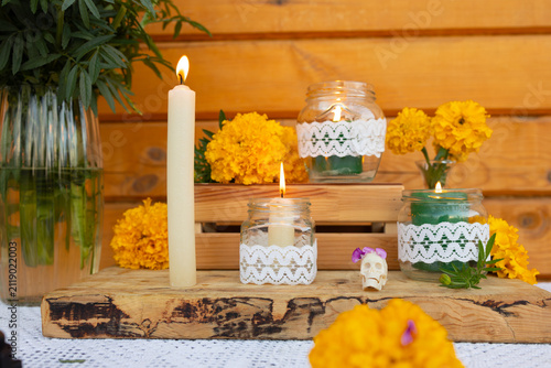 Closeup view of Day of Dead altar featuring lit taper candles in glass jars wrapped with lace, bright marigold blooms, green foliage and rustic wooden table in warm memorial setting