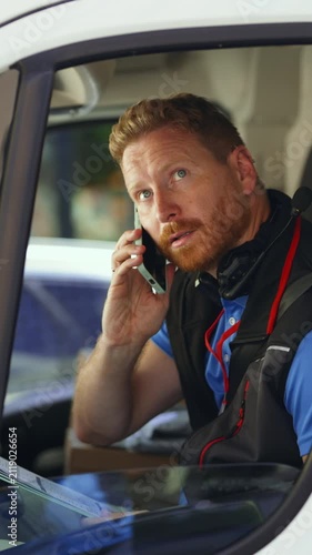 Delivery driver checks a clipboard and talks on a smartphone while seated in a parked van. Courier reviews shipping paperwork during a call, seen through the open window. Vertical video.