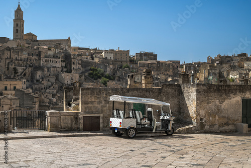 Electric tuk-tuk parked on an ancient stone square in Matera, Italy, with historic Sassi architecture and blue sky in the background. Eco-friendly travel concept
