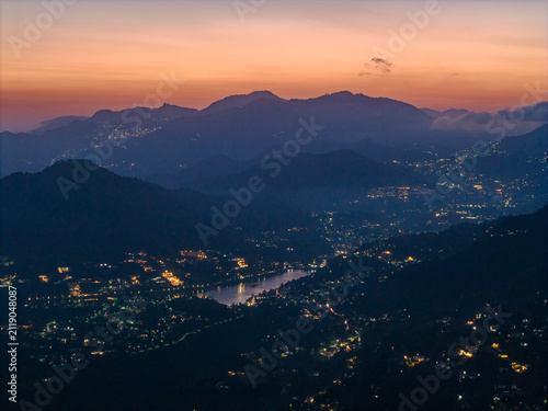 Aerial view of Nainital Lake surrounded by illuminated mountain town buildings and dark forested hills under a soft sunset sky in Nainital, Uttarakhand, India.