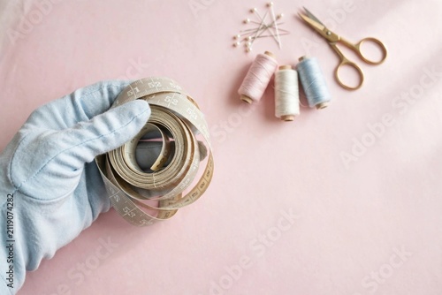 Hand with light blue glove holding coiled measuring tape and sewing supplies on pink surface