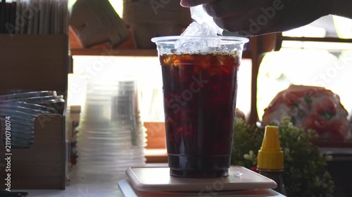 4K Barista adding extra ice cubes to cup of iced black coffee at cozy cafe counter, Concept for iced americano coffee preparation summer refreshment and cafe culture.