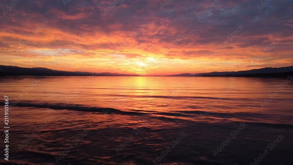 Fototapeta premium Vibrant sunset over calm water with colorful clouds reflecting on the surface, creating a serene atmosphere in a natural landscape at dusk