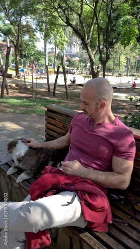 A 50 year old Brazilian man petting his dog in a sunny public square.