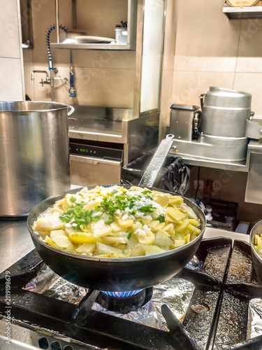 фотографія Sliced potatoes, onions, and green peppers poaching in oil inside a frying pan on a gas stove in a professional kitchen