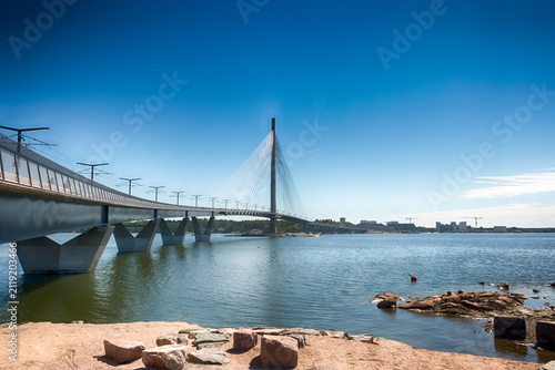 A scenic view of the modern Kruunuvuorensilta cable-stayed bridge over the calm blue sea in Helsinki, Finland, on a bright, cloudless summer day. HDR.