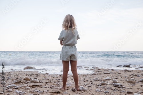 A young girl with blonde hair stands on a pebbly beach looking out at the ocean waves