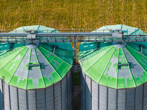 Aerial view of two industrial grain silos with weathered green metal roofs and a connecting catwalk situated in a rural agricultural landscape in Kashubia region, Pomeranian Voivodeshi, Poland.